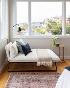 a bed sitting in front of two large windows next to a rug on top of a wooden floor