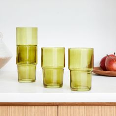 three green glasses sitting on top of a counter next to an apple and a bowl