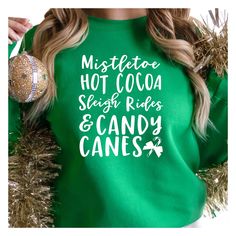 You Can Specify the letter color for the design in the personalization box. Please choose from White, Black, Red, Green, Gold, Silver. If no color is specified then white will be used.  Celebrate the holiday season with this festive "Mistletoe, Hot Cocoa, Sleigh Rides & Candy Canes" design. Available in T-shirt, long sleeve tee, and sweatshirt options, perfect for any winter occasion. Makes a great gift for Christmas, holiday parties, or cozy family gatherings during the festive season. Crafted Winter Name Print Top As Gift, Name Print Tops As Winter Gift, Green Letter Print Top As A Gift, Green Letter Print Top As Gift, Green Letter Print T-shirt For Holiday, Holiday Green T-shirt With Letter Print, Green Tops With Custom Print For Gift, Green Custom Print Top For Gift, Custom Print Green Tops For Gifts