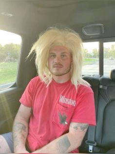 a man with long blonde hair sitting in the back seat of a car wearing a pink shirt