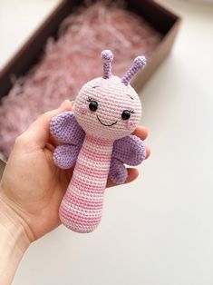 a hand holding a small crocheted toy in front of a box filled with pink stuff