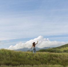 a woman standing on top of a lush green hillside under a blue sky with clouds