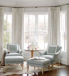 a living room with two chairs and a table in front of large windows that have curtains on them