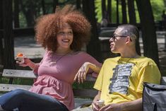 a man and woman sitting on a bench in the park, one holding an orange