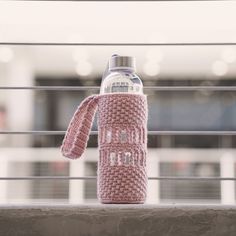 a pink knitted water bottle cover sitting on top of a metal railing next to a building