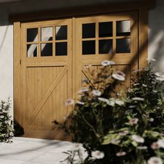 two wooden garage doors in front of a white building with flowers growing on the side