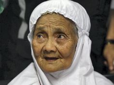 an old woman wearing a white headscarf and looking at the camera with other people around her