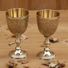 two silver goblets sitting next to each other on a wooden table with leaves