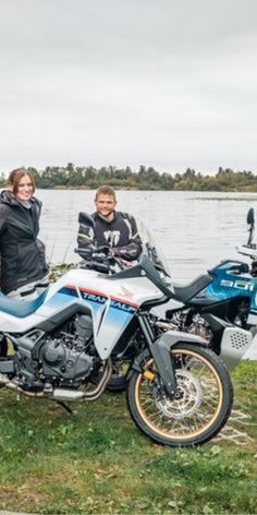 two people standing next to a parked motorcycle near the water on a cloudy day,