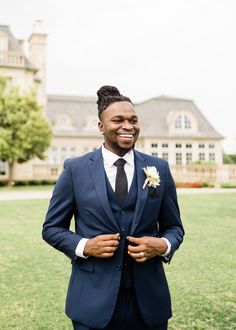 a man in a suit and tie standing in front of a large house smiling at the camera