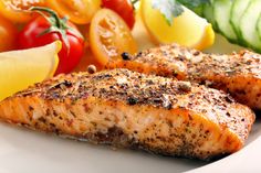 two salmon fillets on a white plate next to some vegetables and lemon wedges