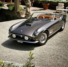 an old fashioned sports car parked in a driveway next to a chair and potted plant