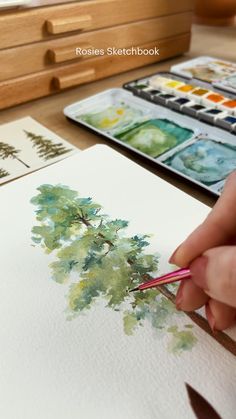 a person is painting with watercolors on paper and using a pencil to draw the tree