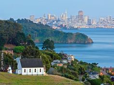 a small white church sits on a hill overlooking the water and city in the distance