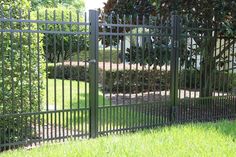 an iron fence in front of a house with green grass and bushes behind it on a sunny day