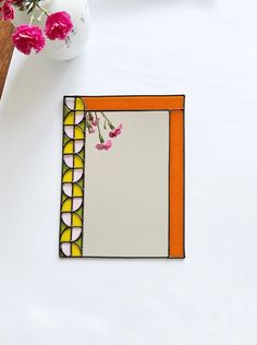 an orange and white frame next to a vase with pink flowers in it on a table