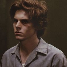 a young man with brown hair wearing a gray shirt and looking at the camera while standing in front of a mirror