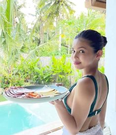 a woman holding a plate with food on it in front of a pool and palm trees