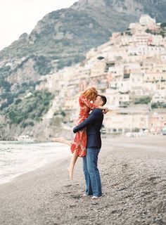 a man holding a woman on the beach in front of a city and mountains behind him