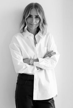 a woman in white shirt and black pants leaning against a wall with her arms crossed