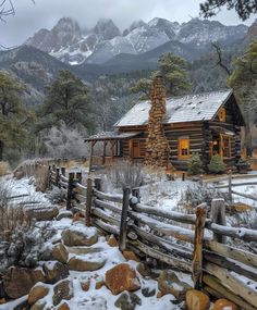 a log cabin in the mountains with snow on the ground and rocks around it, near a wooden fence