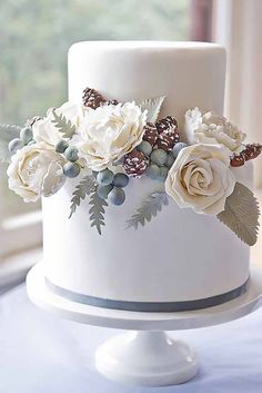 a white wedding cake decorated with flowers and pine cones on a table in front of a window