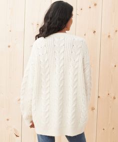 a woman standing in front of a wooden wall wearing a white cable knit sweater and jeans