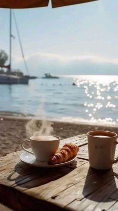 a cup of coffee and some hot dogs on a table at the beach with boats in the water