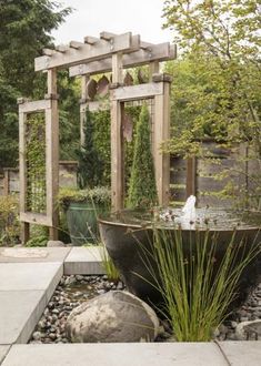 an outdoor fountain surrounded by rocks and plants in the middle of a garden with wooden trelliss