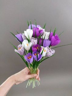 a hand holding a bouquet of purple and white flowers on a gray background with green stems