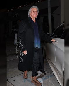 an older man standing next to a car in front of a building at night with his hand on the door handle