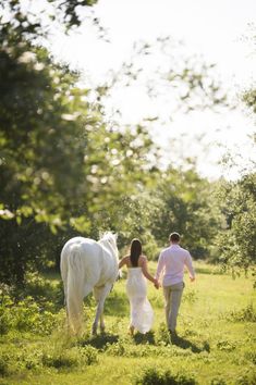 a man and woman are walking with a white horse in the grass near some trees