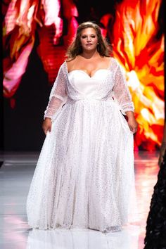 Plus size model wearing a white plus size wedding gown with romantic sheer sleeves and a sweetheart neckline.  The model has her honey blond brown hair half up and down, and she's walking on a white runway with a brilliant bold backdrop. Plus Size Runway, White Wedding Gown, Plus Size Wedding Gowns, White Wedding Gowns