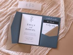 an open pocketfold wedding card on top of a bed next to a note and envelope