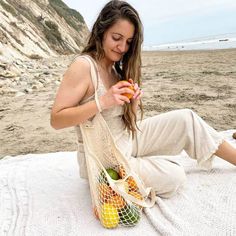 a woman sitting on the beach holding an orange and a bag with fruit in it