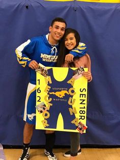 two people standing next to each other in front of a blue and yellow wall holding a sign