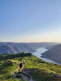 two people walking up a grassy hill towards the water on a sunny day with mountains in the background