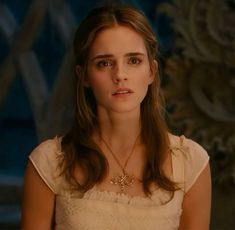 a woman with long hair wearing a white dress and gold necklace in front of a wall