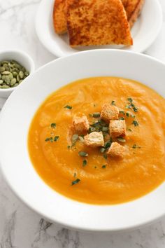 there is a bowl of carrot soup with croutons and bread on the side