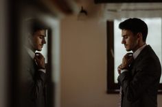 a man adjusting his tie while standing in front of a mirror with another man looking at him