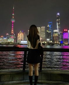 a woman standing on the edge of a pier looking at the city lights in the distance