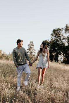 a man and woman holding hands walking through tall grass in an open field with trees behind them