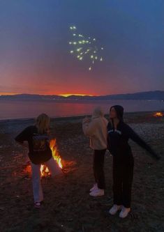 two women standing next to a fire with fireworks in the sky above them at night