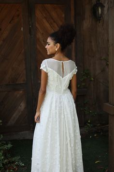 a woman wearing a white dress standing in front of a wooden wall and looking off into the distance