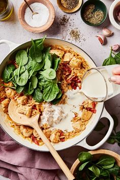 a person pouring milk into a pot filled with pasta and spinach leaves, surrounded by other ingredients