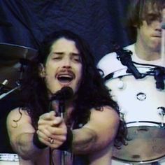 a man with long hair holding a microphone in front of two other men on stage