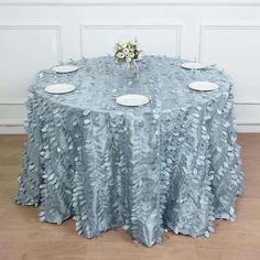 a round table covered in blue cloth with white plates and flowers on the center piece