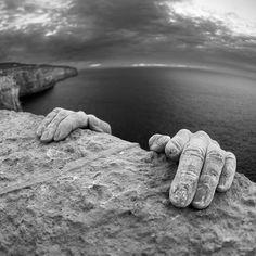 two hands that are on the side of a rock near water and clouds in the sky