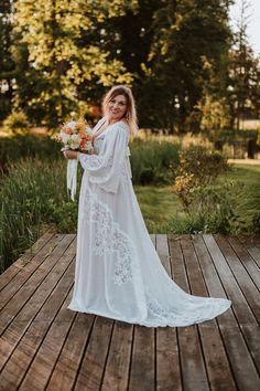 a woman standing on a wooden deck holding a bouquet of flowers and wearing a long white dress