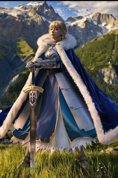 a woman dressed in blue and white standing on top of a grass covered field next to mountains